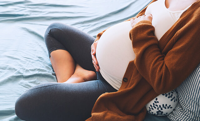 Mulher grávida sentada na cama segurando a barriga, simbolizando as causas e sintomas das varizes relacionados com a gravidez