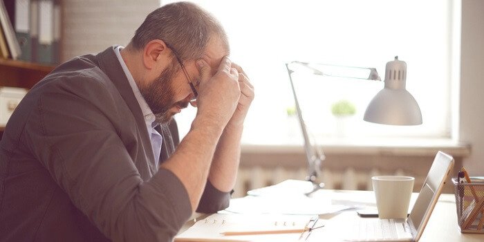 Homem sentado à secretária, com ar cansado e mãos na cabeça, sugerindo stress, um dos fatores que podem contribuir para o surgimento de arritmias.