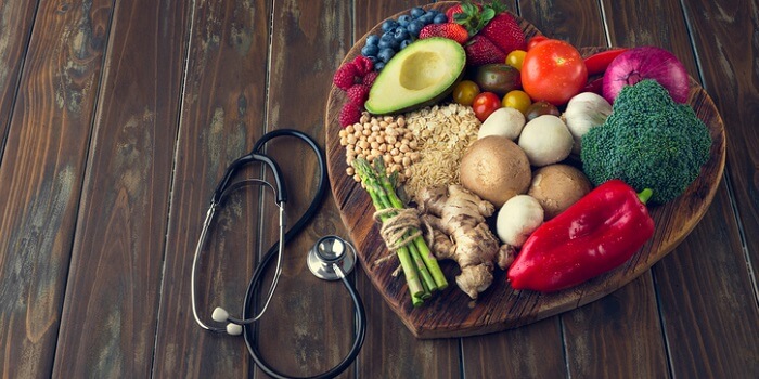 Tábua em forma de coração cheia de alimentos saudáveis, como legumes, frutas, grãos e vegetais, ao lado de um estetoscópio.