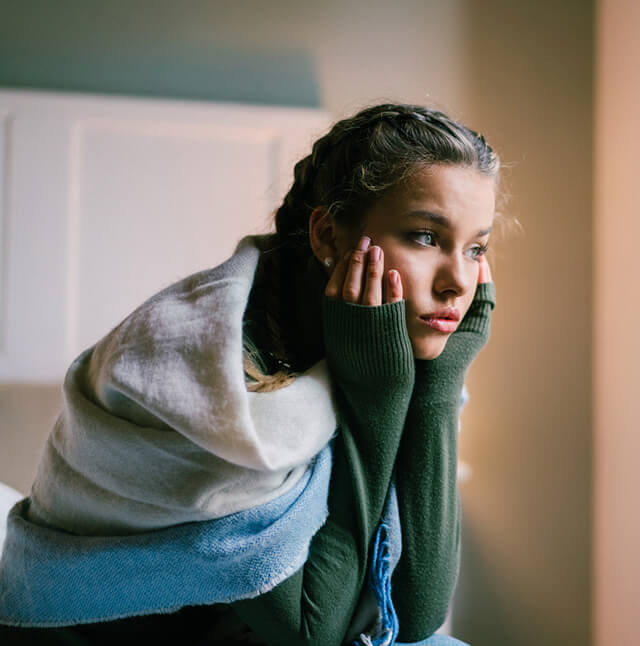 Jovem mulher com uma expressão pensativa e de cobertor nos ombros, representaçõa dos sintomas de ansiedade ou desconforto emocional.