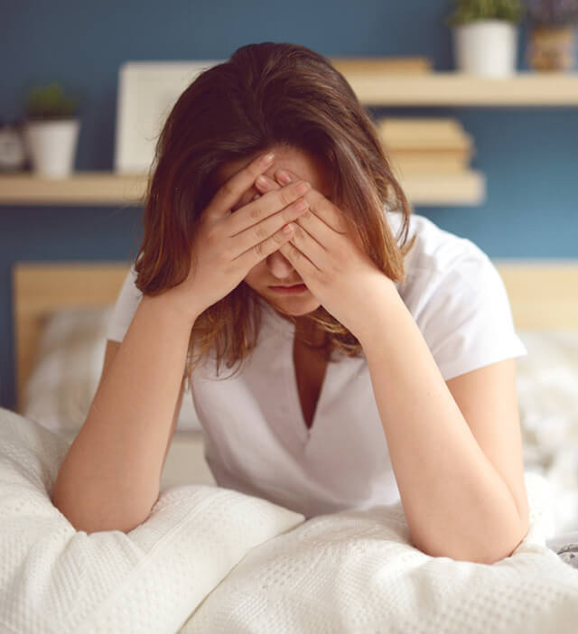 Mulher sentada na cama com as mãos no rosto, revela sinais de exaustão ou tristeza profunda, associados a perturbações depressivas.