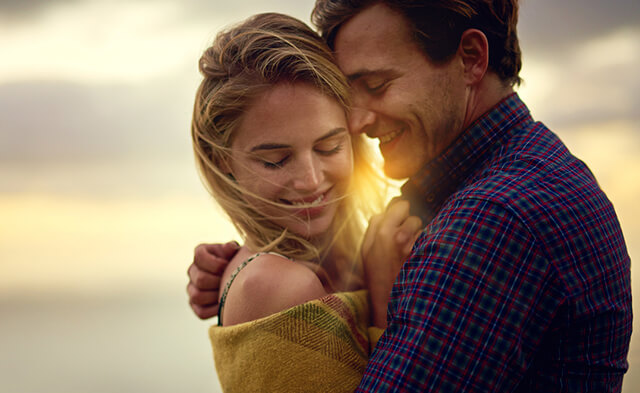 Casal a sorrir e abraçado num ambiente ao ar livre, o que representa a conexão emocional e o bem-estar.