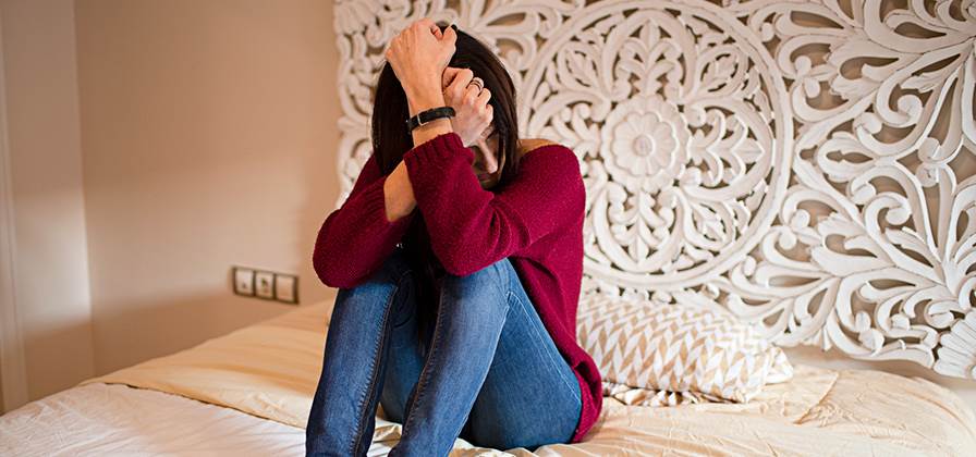 Mulher, sentada na sua cama com ar de profunda tristeza, possível sinal de alguma doença mental.