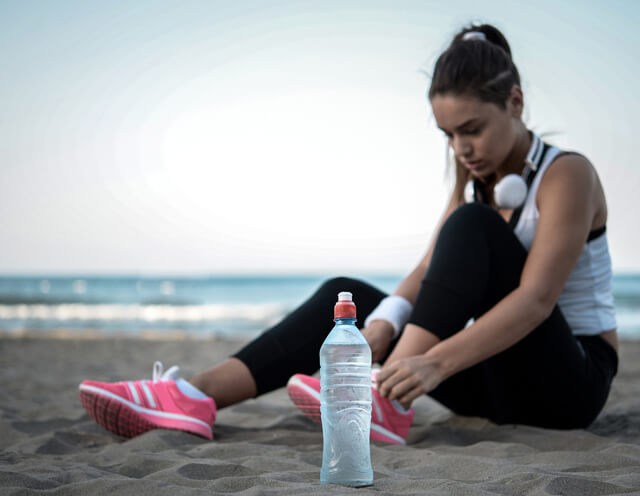 Mulher equipada, a apertar os sapatos, para se preparar para começar a fazer exercício na praia com uma garrafa de água ao lado para estar hidratada.